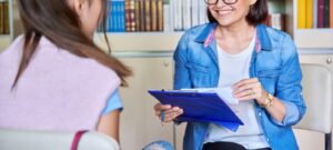 A woman smiles while holding a clipboard and sitting across from a teen. This could symbolize the support a therapist in Boulder, CO can offer for those experiencing screen addiction. Learn more about mindfulness in Boulder, CO and how online therapy in Colorado can offer remote support.
